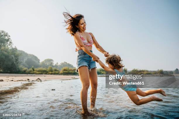 mother whirling daughter around while standing in water at beach - mother and child in water at beach stock pictures, royalty-free photos & images