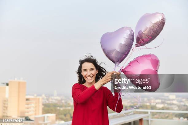 smiling mature woman holding heart shape balloon while standing on building terrace - heart balloon stock pictures, royalty-free photos & images