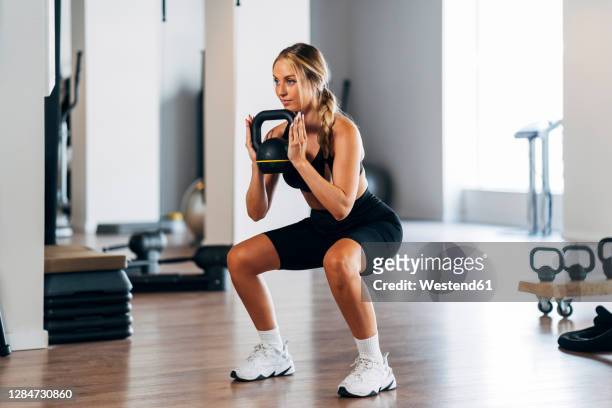 young female athlete lifting kettle bell while crouching in gym - women working out gym stock pictures, royalty-free photos & images