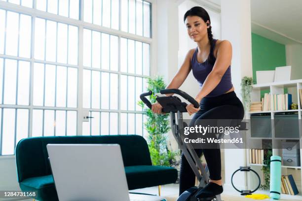 young woman listening music through bluetooth while sitting on exercise bike at home - heimtrainer stock-fotos und bilder