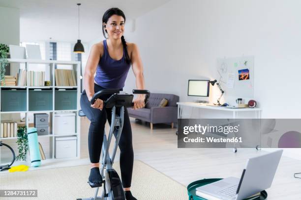 smiling young woman listening music through bluetooth while sitting on exercise bike at home - bluetooth stock-fotos und bilder