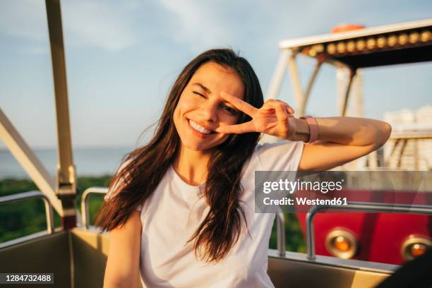 smiling beautiful woman showing peace sign and winking while enjoying ferris wheel ride - friedenszeichen handzeichen stock-fotos und bilder