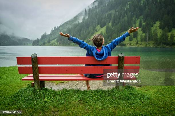 woman sitting on bench with arms outstretched at lakeshore while looking at mountains - landskap stockfoto's en -beelden