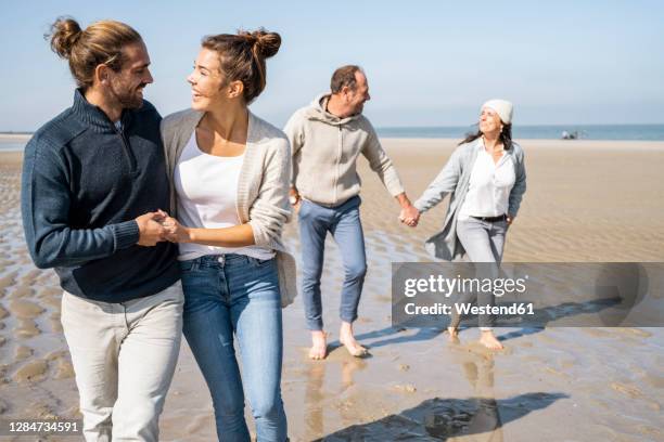 smiling young couple walking with mature couple in background at beach - family trip in laws stock pictures, royalty-free photos & images