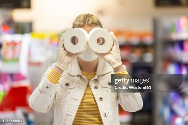 teenage girl wearing protectice mask and gloves holding looking through holes of toilet rolls at supermarket - buying toilet paper stock pictures, royalty-free photos & images