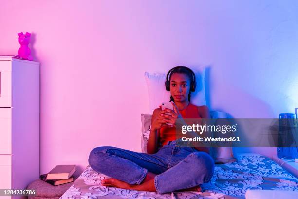 teenage girl sitting on bed using smart phone to listen music through headphone at home - girl in her bed stock pictures, royalty-free photos & images