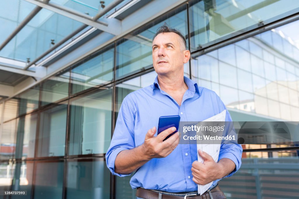 Thoughtful businessman holding smart phone and document while looking away in city during pandemic