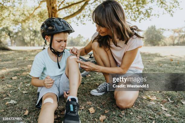 mother putting bandage on son's knee while sitting in public park during sunny day - sports helmet fotografías e imágenes de stock