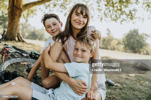 mother embracing sons while sitting in public park - two kids with cycle imagens e fotografias de stock