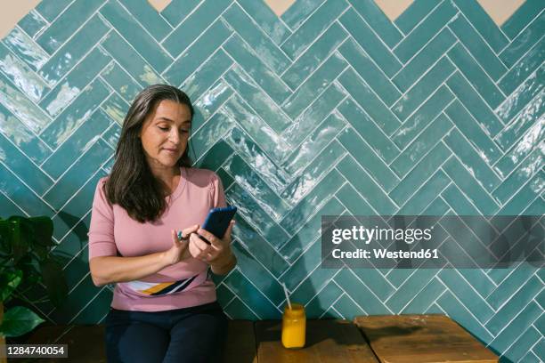 woman using smart phone while sitting with juice against turquoise wall at bar - schwarzes haar stock-fotos und bilder