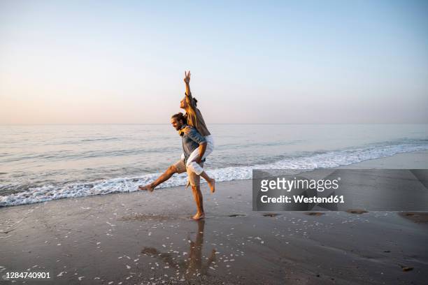 young man giving piggyback to girlfriend on shore at beach against clear sky during sunset - おんぶ ストックフォトと画像