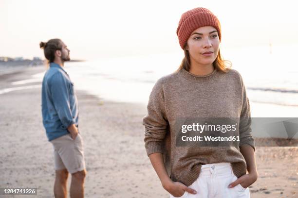 beautiful young woman standing with hands in pockets against boyfriend at beach during sunset - disappointment stock pictures, royalty-free photos & images