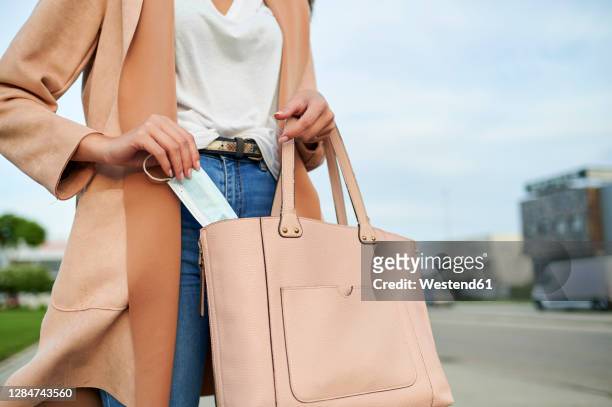 woman taking out protective face mask from purse while standing on street - handtasche stock-fotos und bilder