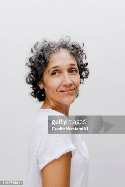 mature woman with short curly hair looking away while standing against white wall - hair woman mature grey hair beauty stockfoto's en -beelden