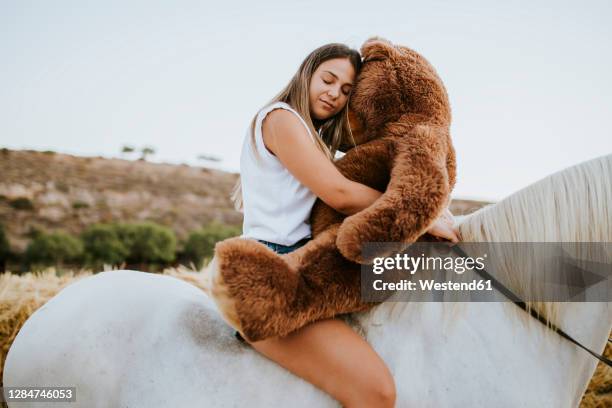 portrait of beautiful young woman embracing large teddy bear while sitting on horseback with closed eyes - mascot stock-fotos und bilder