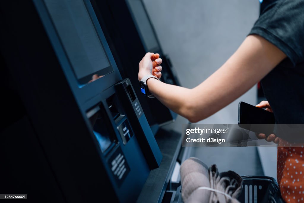 Close up of a young Asian woman using contactless payment via smart watch to pay for her shopping at self-checkout kiosk in a store