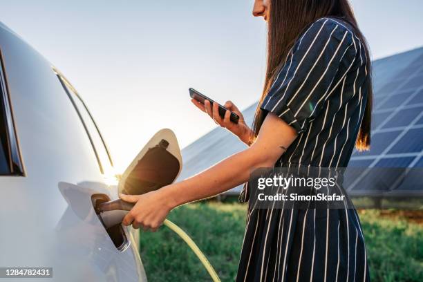 vrouw die elektrische auto en zonnepanelen op achtergrond laadt - opladen stockfoto's en -beelden