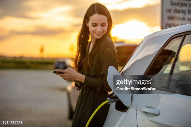 frau wartet auf elektroauto auf dem parkplatz bei sonnenuntergang aufladen - hybrid vehicle stock-fotos und bilder