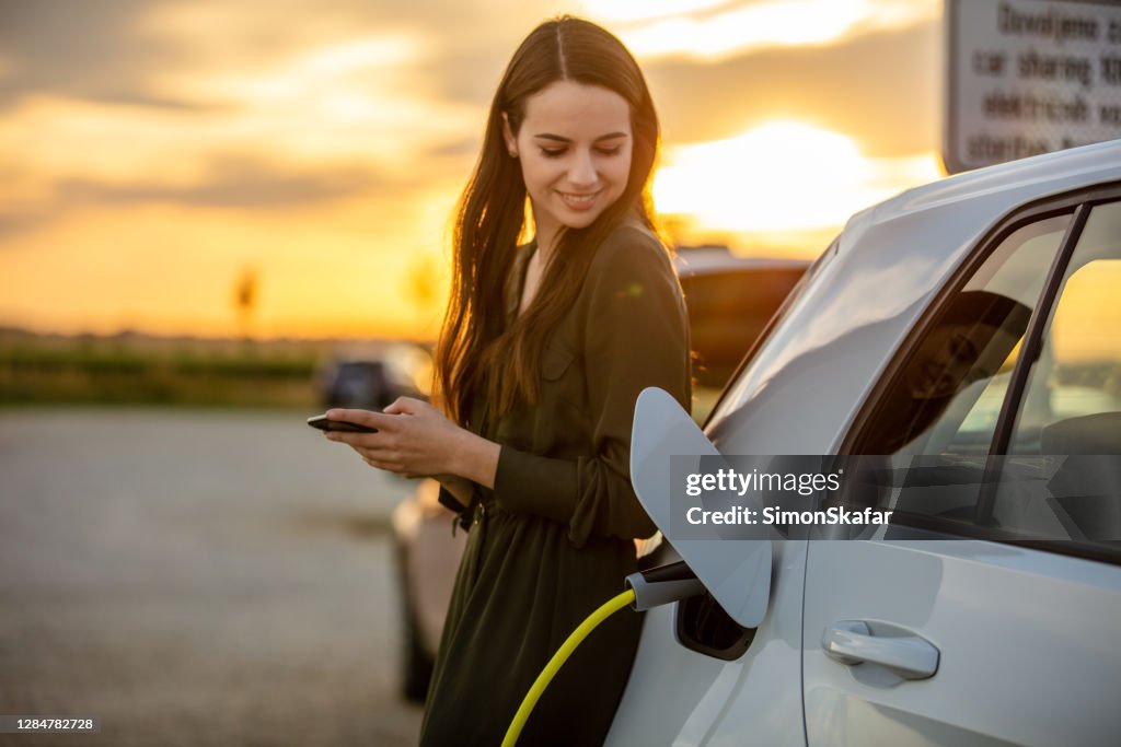 Woman waiting for electric car to charge in the parking lot at sunset
