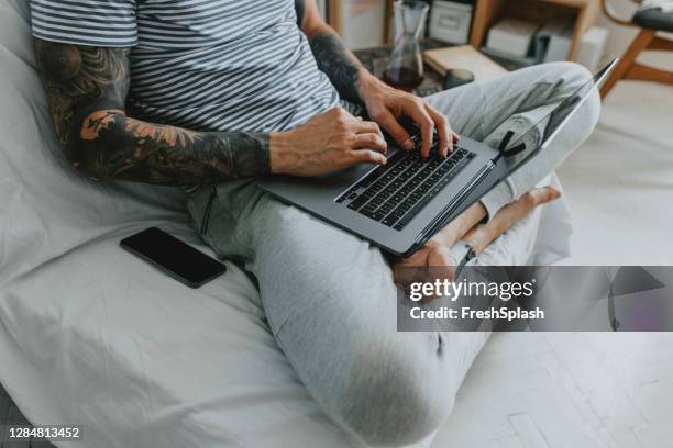 trabajar en casa: anonymous tattooed man haciendo negocios en su computadora portátil desde la comodidad de su sala de estar, adaptándose al nuevo concepto normal - cropped trousers fotografías e imágenes de stock