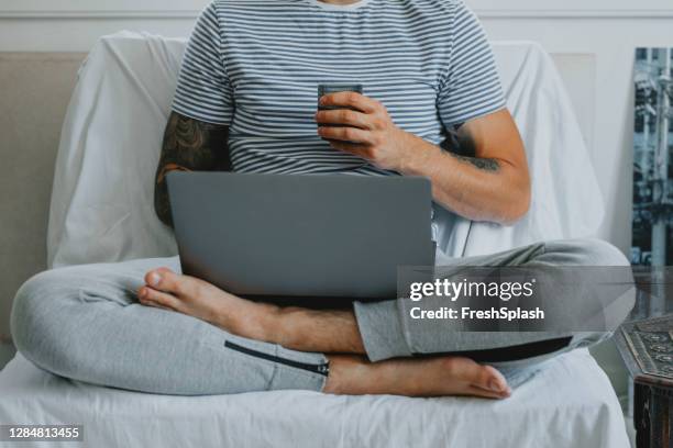 working at home: anonymous young businessman attending an online conference while sitting and drinking coffee in his living room - jogging pants stock pictures, royalty-free photos & images