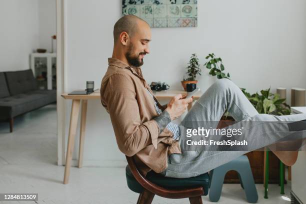微笑的年輕人在社交媒體上與他的朋友交談, 而隨意坐在他的公寓 - jogging pants 個照片及圖片檔