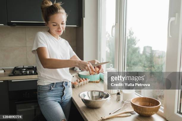 a woman in a white t-shirt and blue jeans prepares an egg omelet for her husband in the kitchen in front of the window - mixing bowl stock pictures, royalty-free photos & images