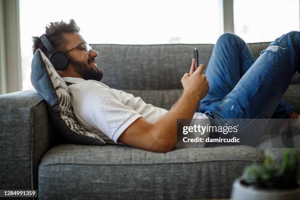 smiling young man with wireless headphones using smart phone on sofa - streaming music stock pictures, royalty-free photos & images