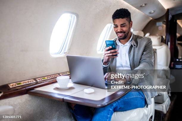 joven hombre de negocios sonríe mientras mira su teléfono inteligente en el jet privado - first class fotografías e imágenes de stock