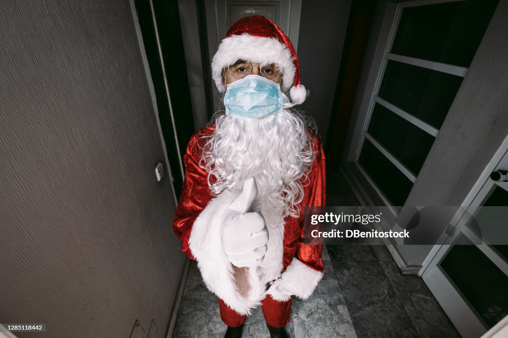 Santa Claus wearing a surgical mask to prevent COVID-19 seen from the peephole of a door, with the thumb of one hand up
