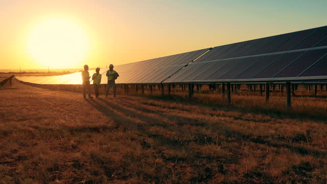 Three solar energy specialists walking through a solar park at sunset. Drone shot