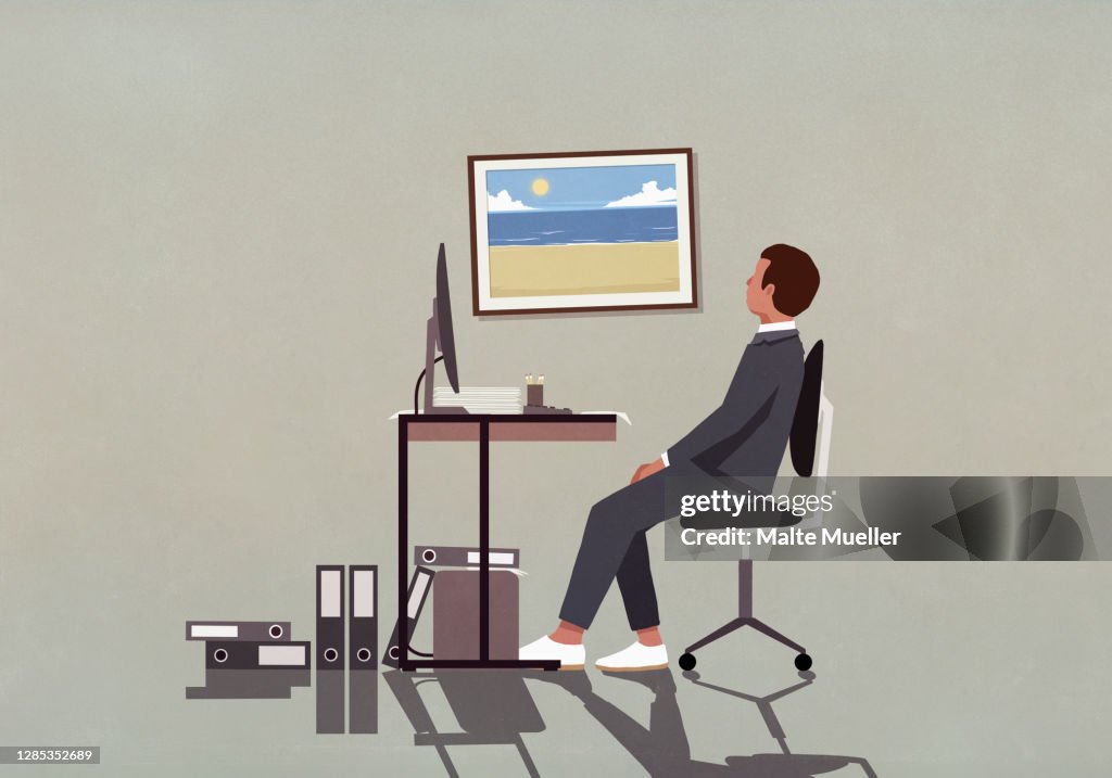 Businessman at office desk looking at photograph of beach on wall