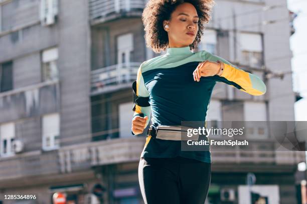 joven mujer afroamericana corriendo en la calle mientras revisa su reloj inteligente - bolsa canguro fotografías e imágenes de stock