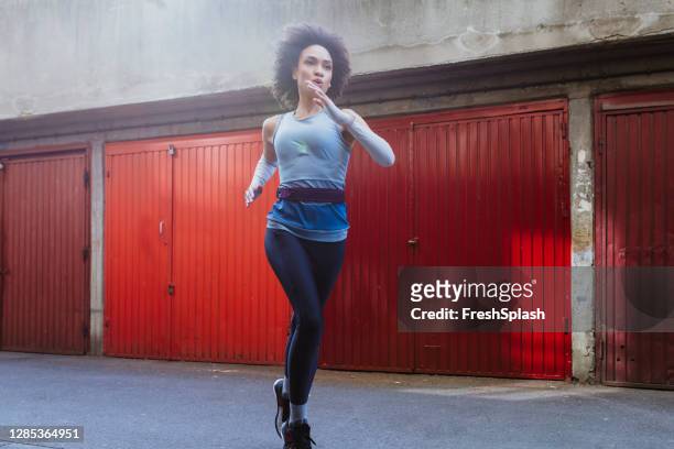 young female afro american athlete running outdoors next to a row of garages - belt stock pictures, royalty-free photos & images