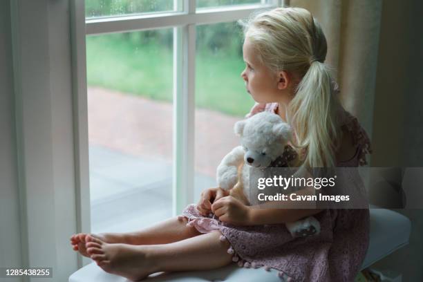 little girl looking sadly out the window watching rain. - kind teddy sitzt stock-fotos und bilder