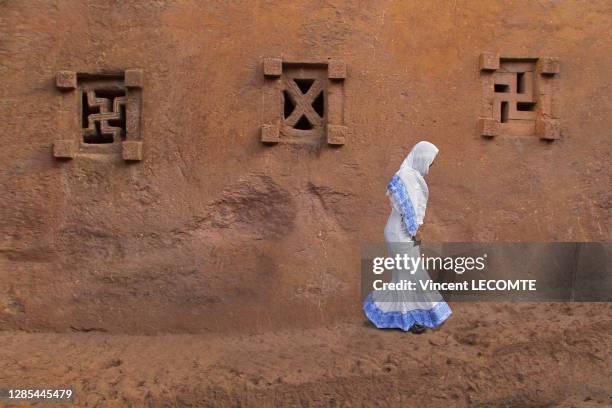 Une femme éthiopienne de profil, vêtue de blanc et bleu marche le long du mur d'une église troglodyte de Lalibela décorée de fenêtres sculptées dans...