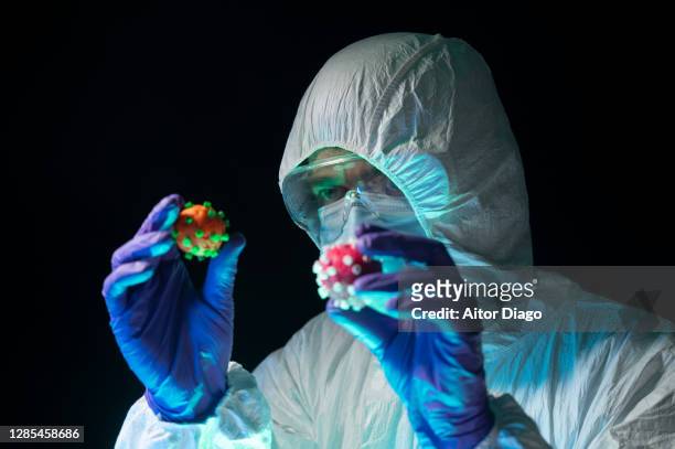 scientist in a protective suit holds and compares two different coronavirus of different color in his hands. creative image. - state of emergency stock pictures, royalty-free photos & images