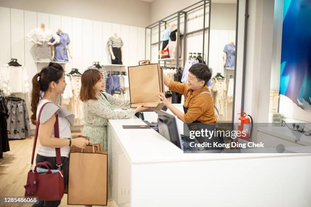 asian chinese cashier hand over the shopping bag to her customers at clothing store counter with smiling face. - clothes shop counter stock pictures, royalty-free photos & images