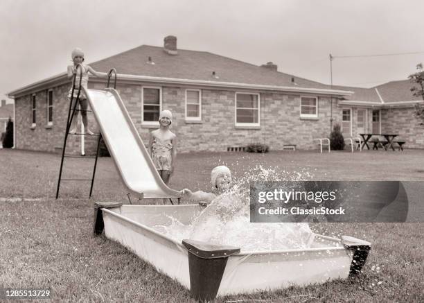 1950s 1960s Three Girls Playing In Backyard Going Down Sliding Board Into Plastic Swimming Pool Splash