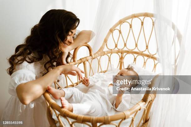 beautiful caucasian mother caressing her baby in rattan crib on white bed linen. - mosquito netting stock pictures, royalty-free photos & images