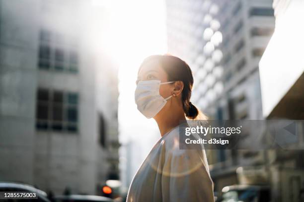 confidence and determined young asian woman with protective face mask standing in the city street, looking away with hope against beautiful lens flare in the fresh morning - nueva-normalidad-concepto fotografías e imágenes de stock