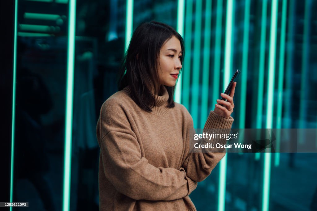 Confident Young Business Woman Using Smartphone Against illuminated light tubes