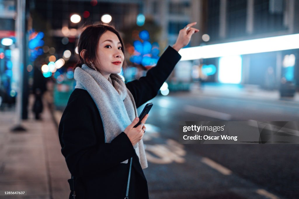 Young Woman With Smartphone Hailing A Taxi In The City At Night