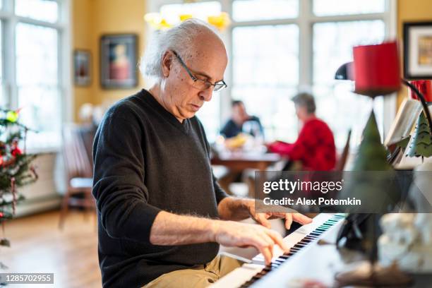 holiday family reunion. the senior 72-years-old silver-hair man, the musician, playing piano for his family who is dinning in the backdrop in the bright spacious living room decorated for christmas. - 60 69 years imagens e fotografias de stock