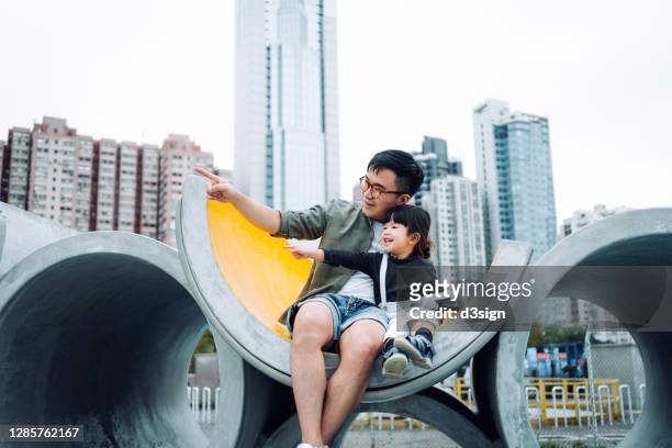 loving asian father with adorable daughter sitting on the concrete water pipe in outdoor playground. smiling joyfully while pointing far away and looking towards sky. enjoying quality father and daughter bonding time, with urban cityscape in background - conveyor belt point of view stockfoto's en -beelden