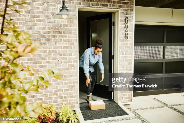 smiling woman picking up package from front porch of home - bulto fotografías e imágenes de stock