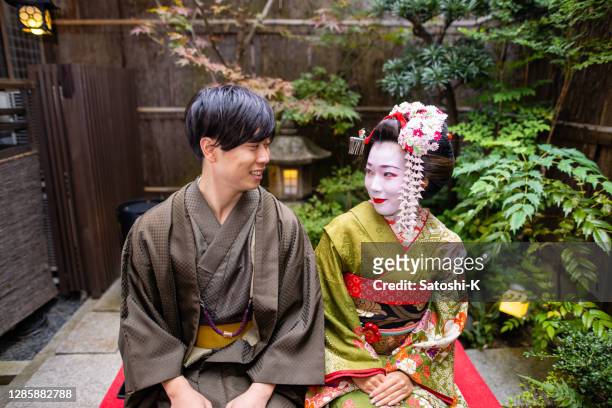 'maiko' (geisha in training) and young man in hakama sitting and looking each other in small japanese garden - three quarter length stock pictures, royalty-free photos & images