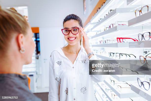smiling young woman trying on red glasses in store - spectacles stock pictures, royalty-free photos & images