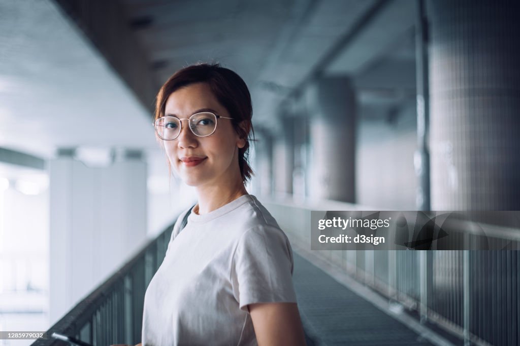 Portrait of confident and successful young Asian businesswoman looking at camera with smile, standing against urban bridge in the city