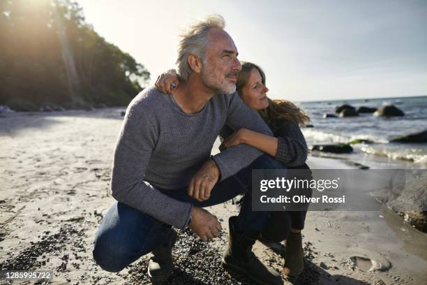 mature couple crouching on the beach looking at view - contemplation couple fotografías e imágenes de stock